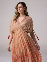 Vestido largo de gasa color salmón con un sutil patrón estampado brillante en tono dorado. Cuenta con escote en V cruzado, cintura imperio elastizada y mangas cortas con tiras ajustables terminadas en borlas.