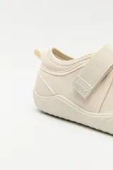 Zapatilla tipo guillermina de estilo barefoot, color beige con detalles en rosa pálido en el talón y tira de ajuste con velcro. Posee suela plana y flexible diseñada para un movimiento natural del pie.