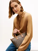 Cartera tote Tommy Hilfiger de acabado texturizado con estampado integral del monograma TH en tonos marrón y negro. Cuenta con dos asas superiores de cuero sintético tostado, bandolera extraíble, cierre de cremallera y herrajes metálicos dorados. Incluye una insignia del monograma en el frente y un detalle de anilla en D.