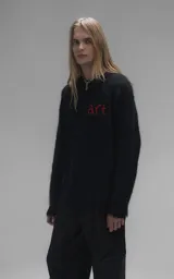 Sweater de punto negro con textura suave, cuello redondo y diseño oversize. Presenta un bordado frontal con la frase 'art SUPPORT' en letras rojas y blancas, y un pequeño detalle de tijeras bordado en la nuca.