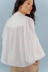 Camisa de algodón color beige con cuello mao, cierre frontal con botones y mangas abullonadas con frunces en los hombros. Presenta costuras verticales decorativas en el frente.