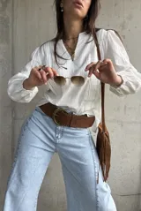 Blusa blanca de mangas largas abullonadas con puños elásticos y detalles de encaje y bordados sutiles. Presenta escote en V con tiras decorativas y diseño holgado.