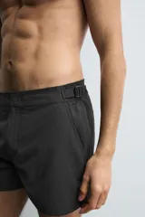 Bañador de hombre color negro, de largo medio, confeccionado en tejido técnico. Presenta cintura elástica ajustable con cordón interior, trabillas laterales, bolsillos frontales y bolsillo trasero con cierre de cremallera.