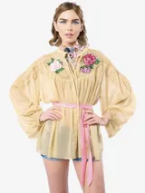 Blusa de gasa color beige con mangas abullonadas, cuello mao y cierre frontal con botones. Presenta aplicaciones de flores bordadas con lentejuelas en el canesú delantero y un lazo de satén rosa en la cintura.
