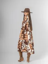 Vestido largo de manga tres cuartos, con cuello camisero y botones frontales. Presenta un estampado abstracto en tonos marrón oscuro, beige y blanco. El calce es amplio y tiene un ruedo con volado.