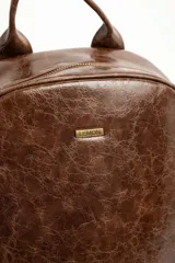 Mochila de cuero sintético marrón con efecto craquelado, asa superior y placa metálica frontal con el logo de la marca.