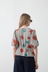 Blusa de silueta relajada con estampado de rayas verticales y motivos florales en tonos naranja y turquesa. Presenta escote en V con lazos frontales regulables, mangas cortas con leve volumen y detalle de frunce bajo el busto que aporta movimiento.