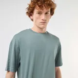 Remera de algodón de corte holgado, color azul grisáceo, con cuello redondo y mangas cortas. Presenta una pequeña etiqueta decorativa en el ruedo inferior.