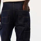Pantalón de jean de mezclilla oscura, corte recto y holgado, con costuras contrastantes en color mostaza/amarillo. Presenta un bolsillo utilitario lateral en la pierna derecha, característico del estilo carpintero.