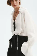 Camisa de lino y viscosa color blanco marfil, de diseño oversized, con cuello alto plisado y volados, mangas largas abullonadas con puños elásticos fruncidos y cordones ajustables en el escote.