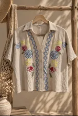 Camisa beige de manga corta con cierre frontal de botones, escote en V y bordados artesanales de flores en tonos azules, amarillos y rojos, acompañados de patrones geométricos.