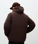 Campera tipo parka color marrón con capucha estructurada, cierre frontal y bolsillo vertical con cierre en el pecho. Presenta detalles de estampado utilitario en el frente y recortes en el cuerpo.