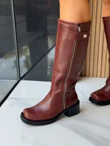 Bota de caña alta estilo biker, confeccionada en cuero color chocolate. Presenta un cierre metálico lateral que recorre toda la caña, estribo y cadena decorativa desmontables, y taco cuadrado con plataforma.