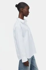 Camisa blanca oversized de manga larga, con cuello clásico, cierre frontal de botones, un bolsillo aplicado en el pecho y dobladillo redondeado.