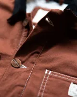 Campera estilo workwear de Denali, confeccionada en canvas resistente color marrón. Presenta interior forrado en corderito sherpa, cuello de pana negro, cierre frontal con botones metálicos color cobre y cuatro bolsillos frontales con costuras reforzadas en contraste.