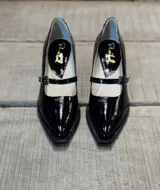 Zapato mocasín de charol negro, con punta fina, tira sobre el empeine y hebilla metálica.