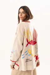 Saco tipo cardigan oversize de punto color beige, con diseño temático marino que incluye bordados de corales, peces, estrellas de mar y tortugas. Presenta detalles de galón picot en color rojo en los bordes y puños, cierre frontal con botones al tono y corte holgado.