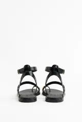 Sandalias planas de cuero color negro, con tiras finas, sujetador para el dedo y detalle de anilla metálica en la puntera. Cuenta con tira ajustable alrededor del tobillo con hebilla plateada.