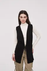 Chaleco largo sin mangas de tejido de punto color negro, con capucha y lazo ajustable en la cintura. Presenta bolsillos frontales y diseño abierto.