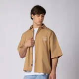 Camisa de manga corta color tostado con corte boxy, cuello camisero, cierre frontal de botones y bordado pequeño en el pecho que dice "VCP CNPT 333".