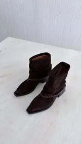 Bota de caña media confeccionada en cuero gamuzado color marrón oscuro, con diseño de caña arrugada y detalle de costura trenzada en la base. Cuenta con un taco cuadrado de madera y punta cuadrada.