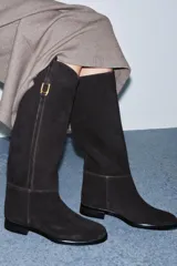Bota de caña alta confeccionada en serraje, con diseño de estilo hípico. Presenta detalle de pespuntes verticales, tira decorativa con hebilla metálica en la parte superior de la caña y puntera redonda. Cuenta con suela plana de 2,5 cm y sistema de calce tipo pull-on.