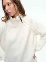 Sweater de tejido de punto acanalado en color blanco, con cuello alto y cierre metálico diagonal en el hombro. Presenta mangas largas con diseño raglán y corte holgado.