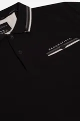 Remera polo azul marino de manga corta, con cuello y puños a rayas blancas y verde militar. Presenta un bolsillo en el pecho con detalle de franja a rayas y el logo de la marca "Brooksfield" bordado en dorado.