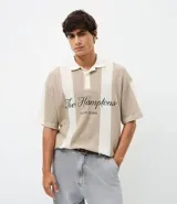 Remera polo de punto con diseño de rayas verticales en tonos beige y blanco. Presenta un corte oversized, cuello clásico con abertura de botones y bordado frontal con la inscripción The Hamptons.