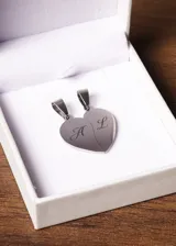 Conjunto de dos dijes de acero quirúrgico con forma de corazón, diseñados para compartir en pareja. Cada dije tiene una inicial grabada, uno con la letra "A" y el otro con la letra "L".