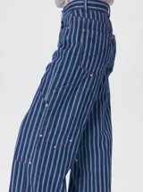 Pantalón de tiro medio y pierna ancha confeccionado en denim de algodón grueso. Presenta un diseño a rayas verticales en tonos azul y celeste, con costuras en contraste color rojo, bolsillos laterales tipo cargo y remaches metálicos.