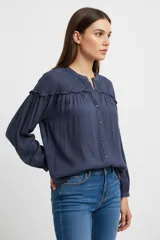 Camisa azul de corte holgado con cuello a la base, cierre frontal con botones y mangas largas abullonadas. Presenta detalles de plisado en los hombros y un delicado volado horizontal que recorre el pecho y la espalda.