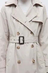 Trench coat de corte clásico en color beige, con cierre frontal de doble botonadura, solapas amplias y cinturón ajustable con hebilla. Incluye capucha desmontable y detalles de trabillas en los puños.