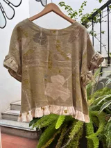 Top de lino con diseño de impresión botánica (ecoprint) en tonos verdes y tierra. Presenta un corte holgado, mangas cortas con volados y un volado decorativo en el ruedo.
