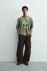 Camiseta de corte relajado con cuello redondo y manga corta. Presenta un efecto de lavado desgastado en tonos marrones y un estampado frontal de una cabeza de alienígena en color verde vibrante.
