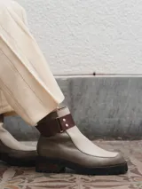 Bota corta de cuero color gris y beige, con detalle de correa marrón con hebilla y tachas plateadas. Tiene suela de goma negra y taco de madera.