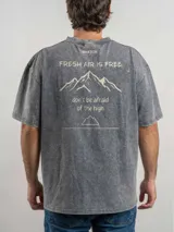 Remera de corte oversize color verde con efecto lavado, manga corta y cuello redondo. Presenta un estampado gráfico en la espalda con el diseño de una montaña y la frase 'Fresh air is free, don't be afraid of the high'.