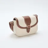 Bolso bandolera de diseño estructurado, confeccionado en lona color beige con detalles de cuero sintético marrón en los bordes y la solapa frontal. Cuenta con una hebilla decorativa metálica, costuras a la vista en color blanco y correa ajustable.