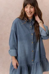 Vestido camisero corto de denim liviano azul con efecto desgastado, de corte oversize, con cuello camisero, botonadura frontal, bolsillo plaqué en el pecho, mangas largas y volado fruncido en el ruedo.