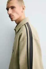 Sudadera de corte boxy fit color verde oliva, con cuello solapa y abertura frontal. Presenta mangas largas con bandas laterales negras a contraste y bordado frontal con texto en color negro.