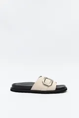 Sandalia slide color beige con hebilla metálica plateada y suela negra.