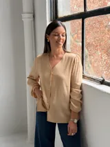Blusa de color tostado con cuello en V, cierre frontal con botones y mangas largas con puños elásticos. Presenta un diseño de corte recto con bolsillos laterales integrados.