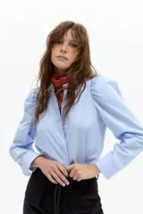 Camisa celeste de popelina de algodón con cuello mao, cierre frontal abotonado con botones de nácar y hombros levemente fruncidos con mangas largas abullonadas.