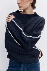 Jersey de punto color marrón chocolate, de cuello alto y corte holgado. Presenta un diseño acanalado y un detalle de costura decorativa en color blanco que recorre la parte superior de las mangas.