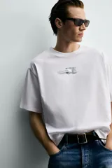 Camiseta de corte boxy fit, color blanco, con cuello redondo y manga corta. Presenta una aplicación de etiquetas decorativas a contraste en el centro del pecho.