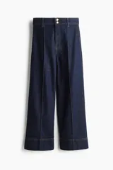 Jean ancho tobillero de denim azul oscuro con leve stretch, tiro alto y cierre frontal con dos botones metálicos. Presenta perneras amplias con pliegues cosidos al frente y dobladillos anchos.