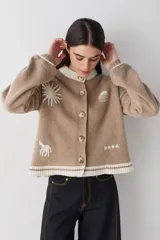 Cardigan de lana color crema con cuello redondo, cierre frontal de botones y mangas largas con puños ajustados. Presenta bordados decorativos en el frente que incluyen un sol, olas y estrellas, con detalles en azul en el cuello y el ruedo.