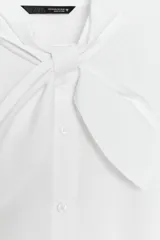 Camisa de popelín de algodón con cuello subido y detalle de aplicación tipo pañuelo anudado al cuello. Presenta manga larga con puños clásicos y cierre frontal mediante botones.