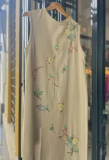 Vestido midi sin mangas color beige, confeccionado en lino de algodón, con delicado bordado artesanal de flores coloridas dispersas por la prenda y tajo frontal.