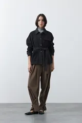Sobrecamisa de jean color negro con silueta relajada, cuello camisero, cierre frontal con botones metálicos, bolsillos frontales y cinturón ajustable en la cintura.
